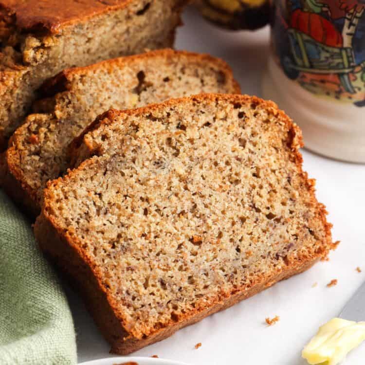 Close-up of moist and tender slices of healthy banana bread on a white surface, with a green napkin, ripe bananas in the background, and a colorful fall-themed coffee mug. A buttered knife and a partially eaten slice are visible in the foreground.
