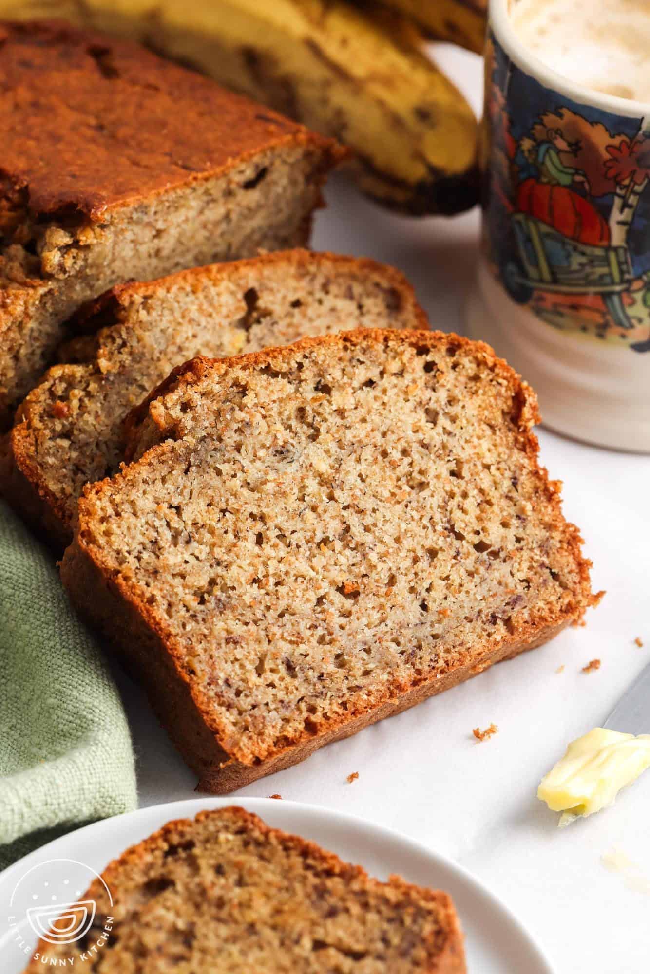 Close-up of moist and tender slices of healthy banana bread on a white surface, with a green napkin, ripe bananas in the background, and a colorful fall-themed coffee mug. A buttered knife and a partially eaten slice are visible in the foreground.