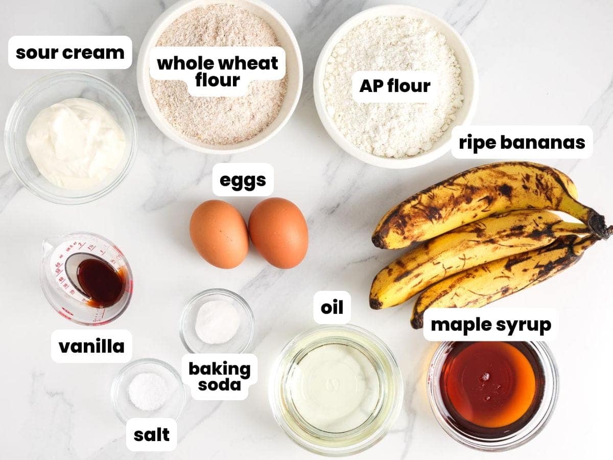 Overhead view of ingredients for healthy banana bread, including ripe bananas, whole wheat flour, all-purpose flour, sour cream, eggs, maple syrup, oil, vanilla, baking soda, and salt.