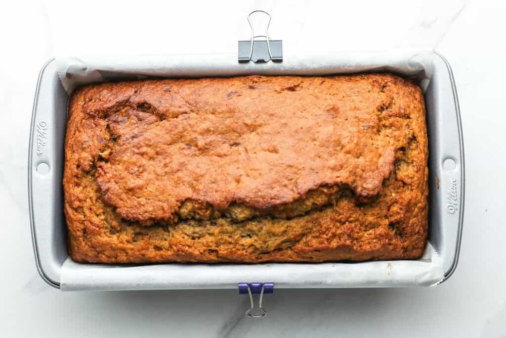 Freshly baked healthy banana bread in a parchment-lined loaf pan, golden brown with a cracked top.