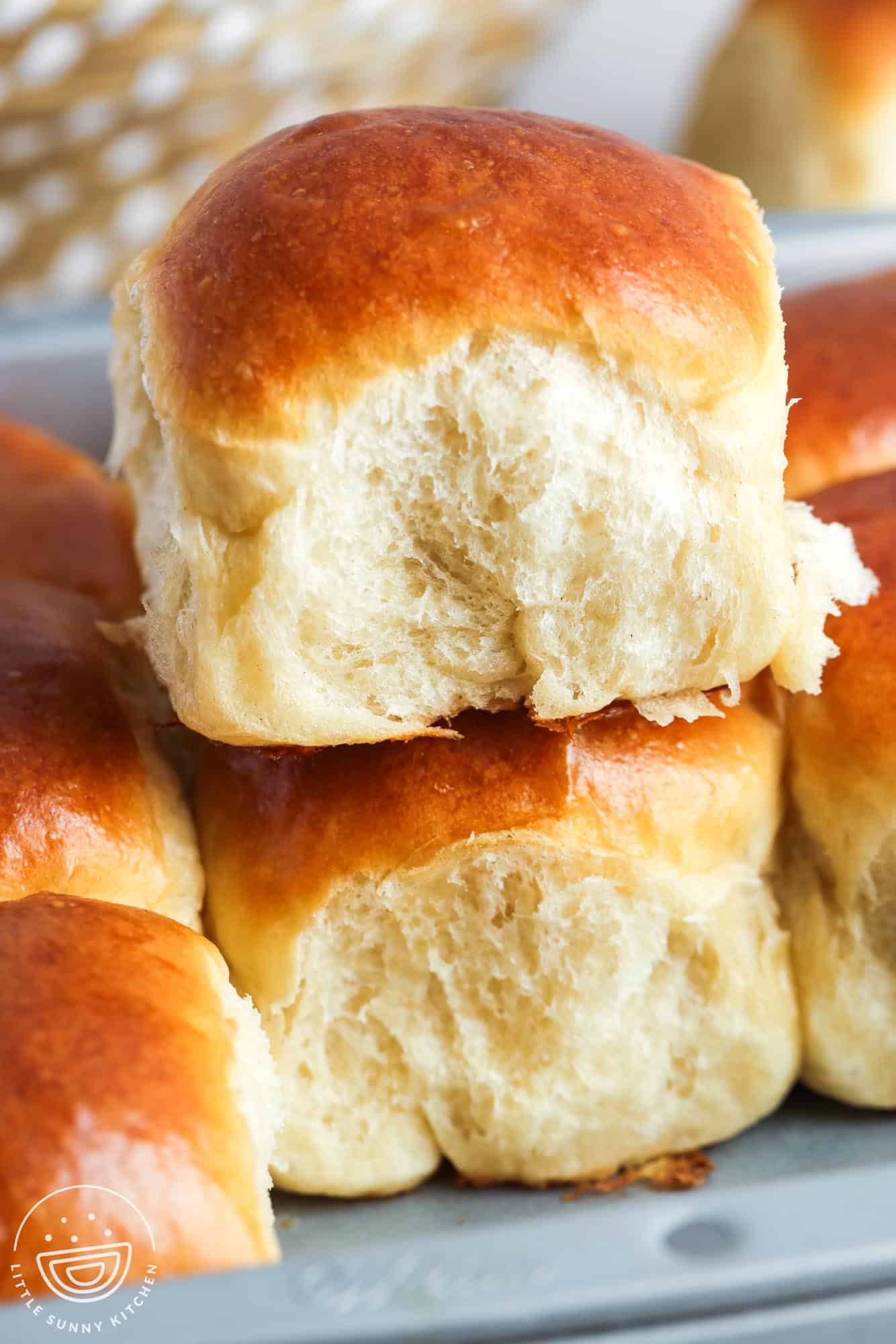 Fluffy golden-brown dinner rolls stacked on a baking tray. The top roll is pulled slightly apart, revealing its soft, airy interior. In the background, more rolls and part of a wicker basket can be seen. 