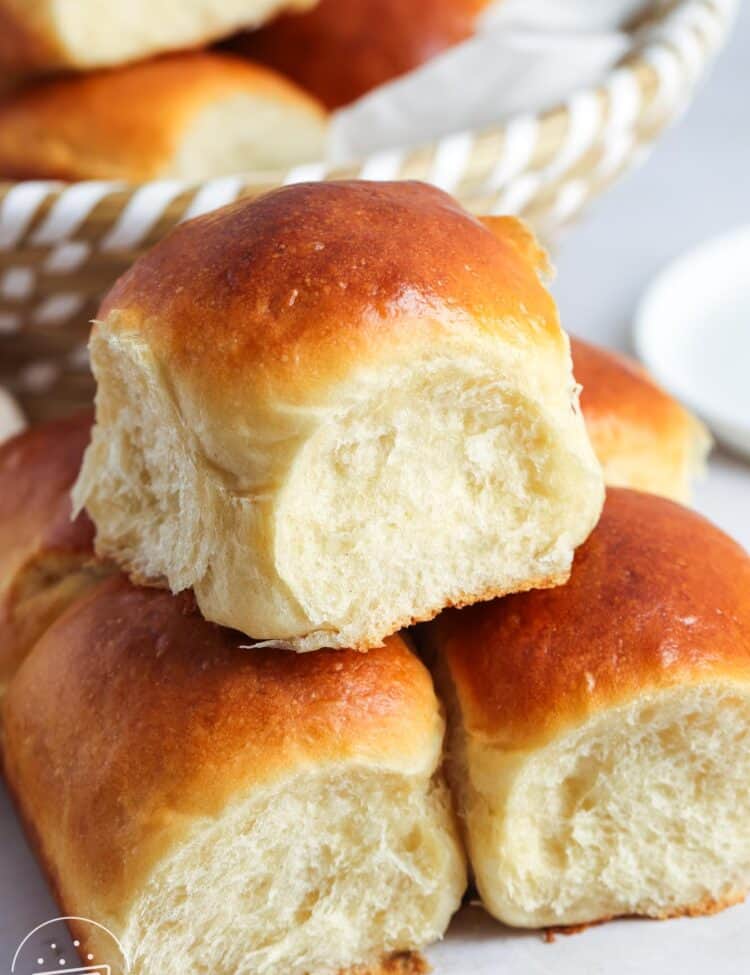 A close-up of soft, golden-brown dinner rolls stacked on a white surface, with a basket of more rolls in the background. The rolls have a fluffy, pillowy interior and a shiny, perfectly baked top. A small circular logo in the bottom left corner reads "Little Sunny Kitchen."