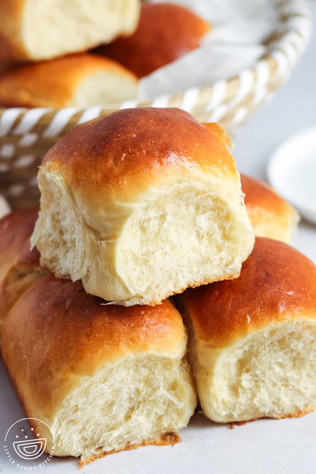 A close-up of soft, golden-brown dinner rolls stacked on a white surface, with a basket of more rolls in the background. The rolls have a fluffy, pillowy interior and a shiny, perfectly baked top. A small circular logo in the bottom left corner reads "Little Sunny Kitchen."