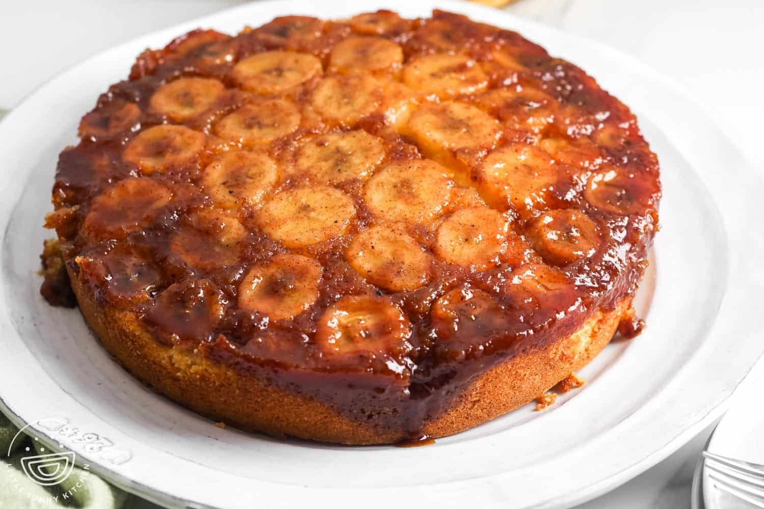 Whole banana upside down cake on a white platter with a slice cut out, showing caramelized banana topping.