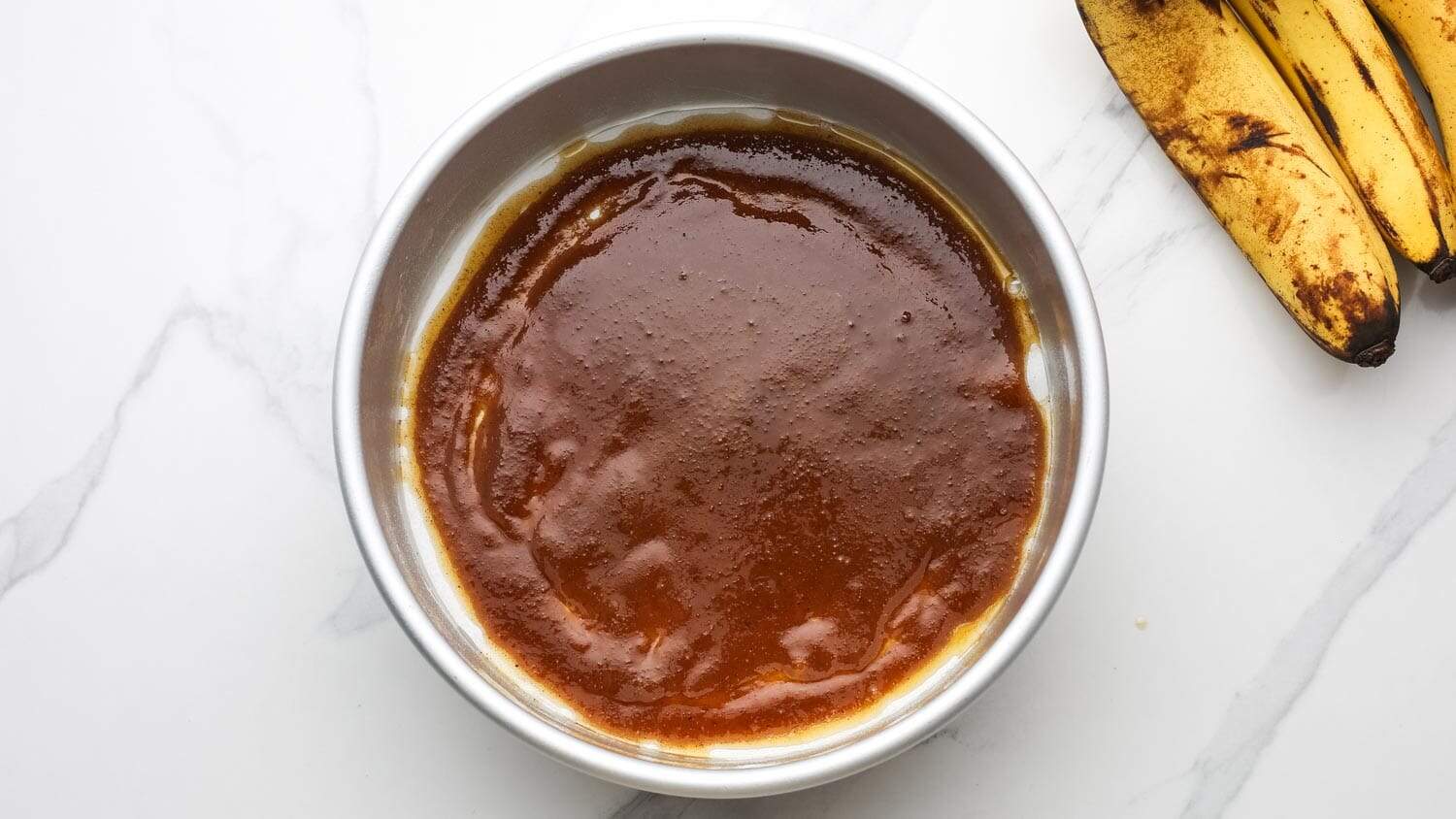 Caramel topping poured into the bottom of a round cake pan with bananas nearby.