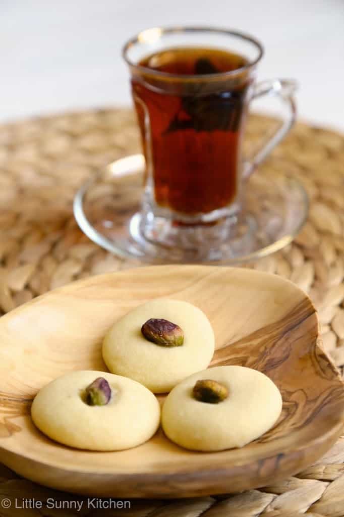 3 ghraybeh cookies on a wooden small plate, with a cup of arabic tea in the background