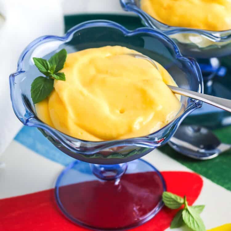 A blue glass dessert cup filled with creamy mango mousse, garnished with fresh mint, with text overlay that reads "3 ingredient Mango Mousse" and "Little Sunny Kitchen."