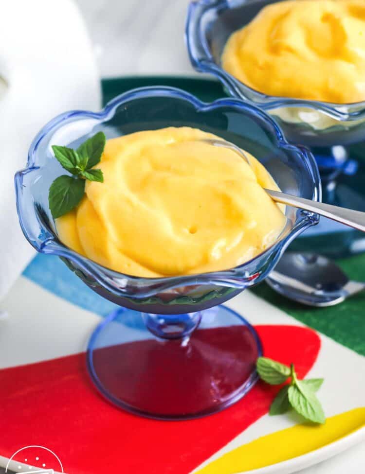 A blue glass dessert cup filled with creamy mango mousse, garnished with fresh mint, with text overlay that reads "3 ingredient Mango Mousse" and "Little Sunny Kitchen."