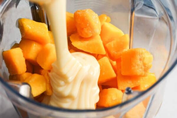 Frozen mango chunks in a blender with sweetened condensed milk being poured on top