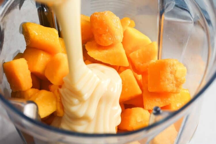 Frozen mango chunks in a blender with sweetened condensed milk being poured on top