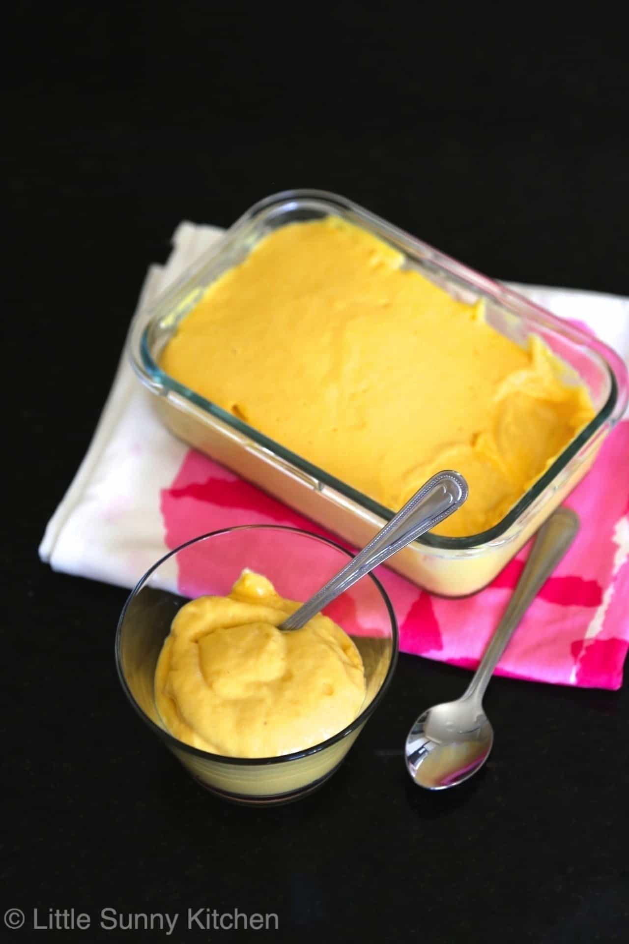 A glass container filled with mango mousse and a bowl of mango mousse with a silver spoon in it.