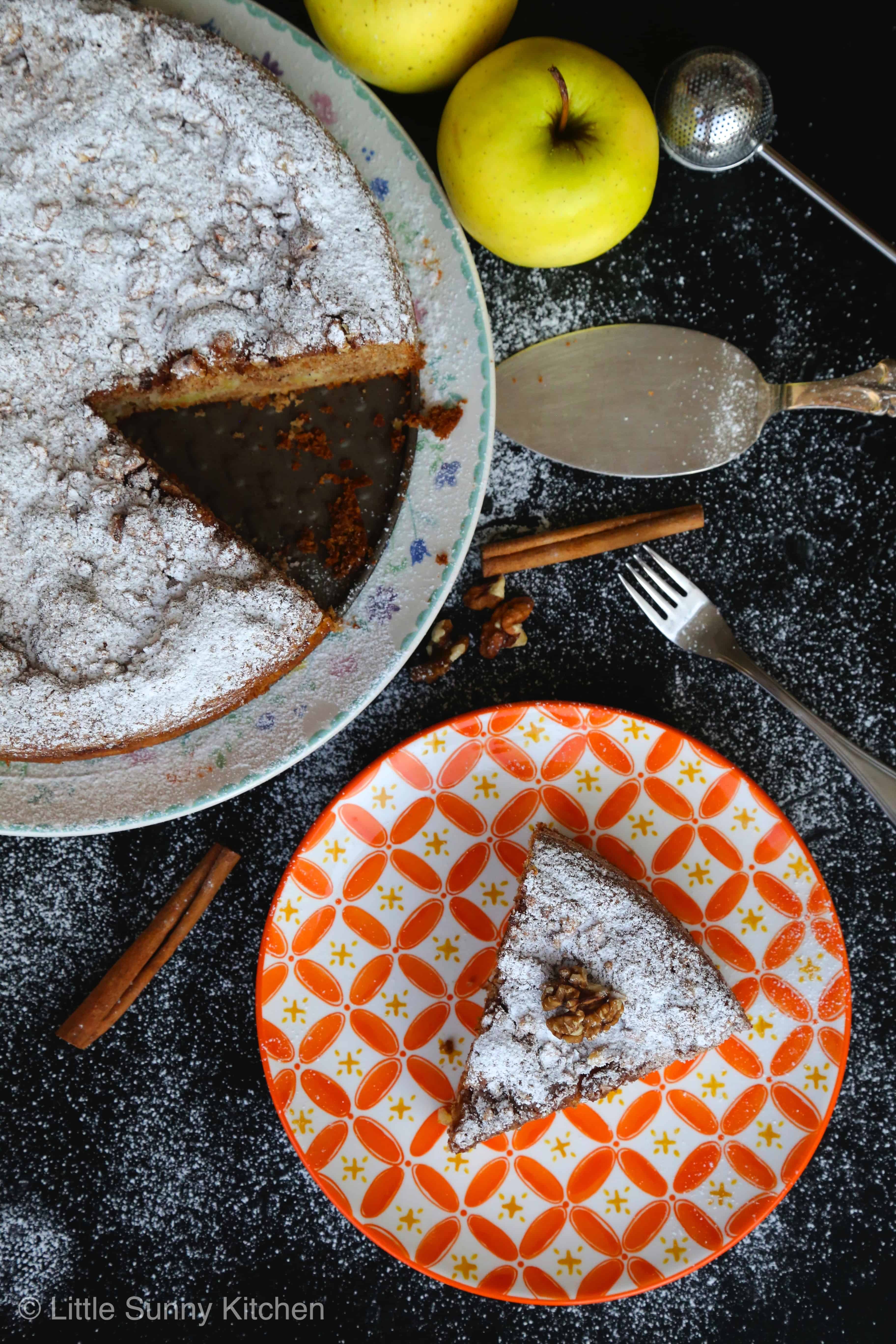 Top view of an apple Sharlotka cake slice served in an orange patterned plate with the cake, cinnamon sticks, and apples on the side. 