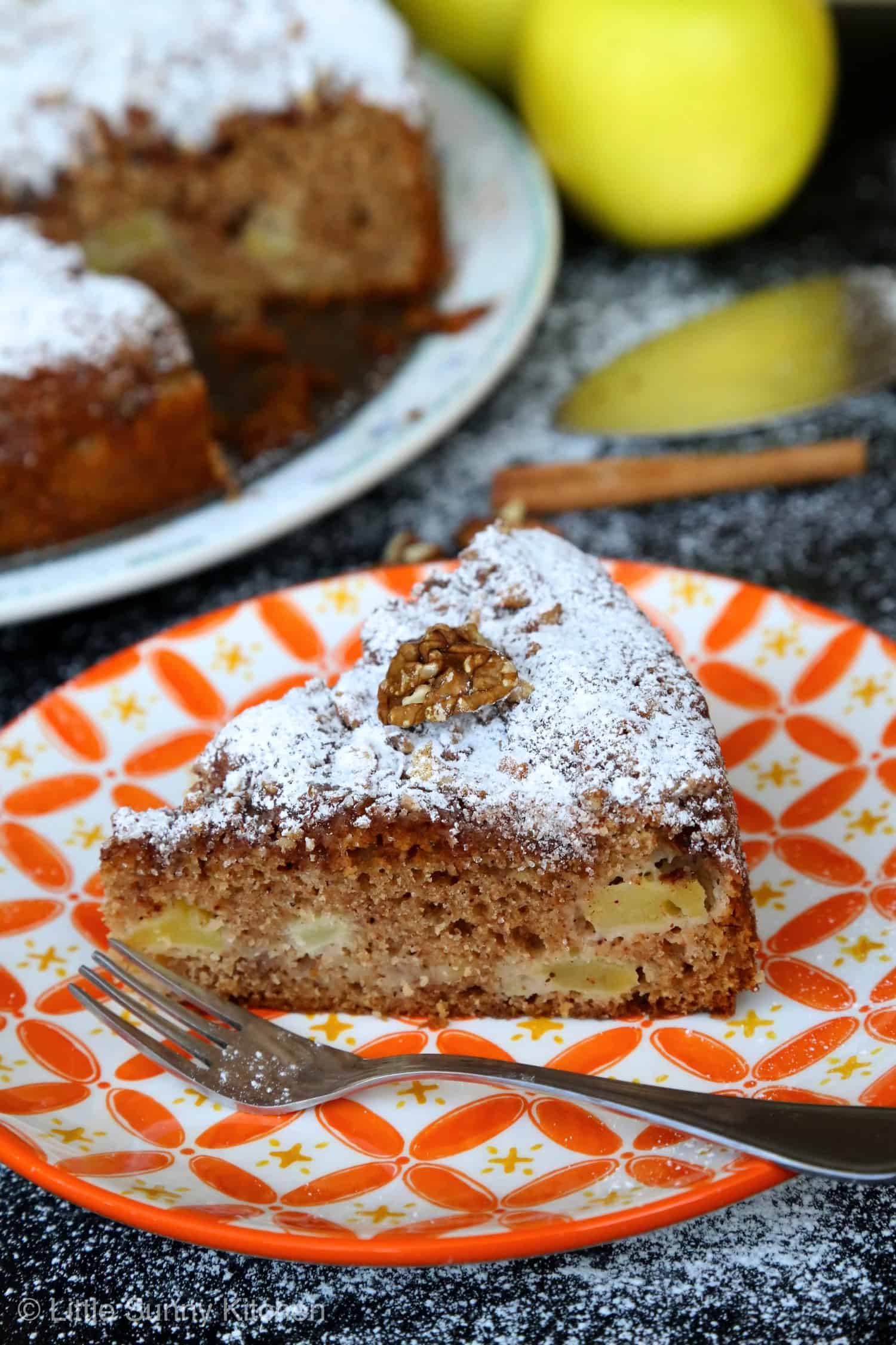 A slice of apple Sharlotka cake topped with powdered sugar served in an orange patterned plate and a fork.