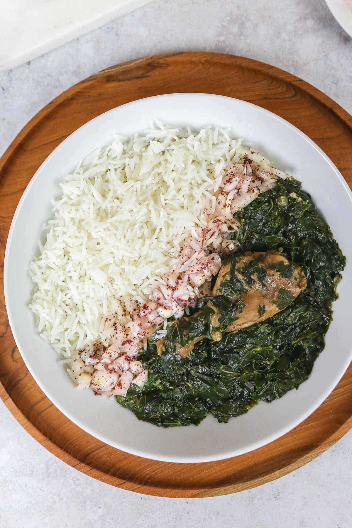 Une assiette blanche avec de la Molokhia, un pilon de poulet, du riz blanc cuit et un mélange de sumac à l'oignon. Posé sur un plateau en bois.