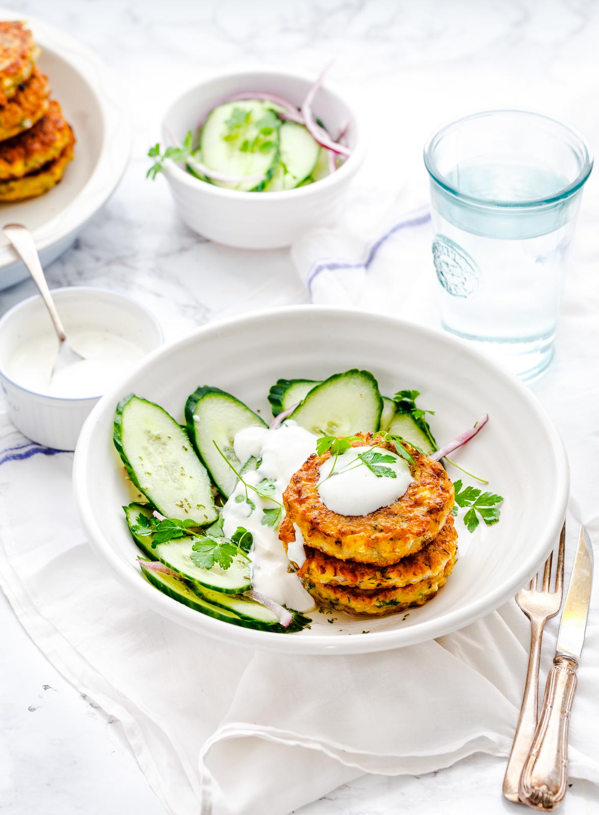 Corn fritters in a white plate, with a cucumber salad, and yogurt sauce.