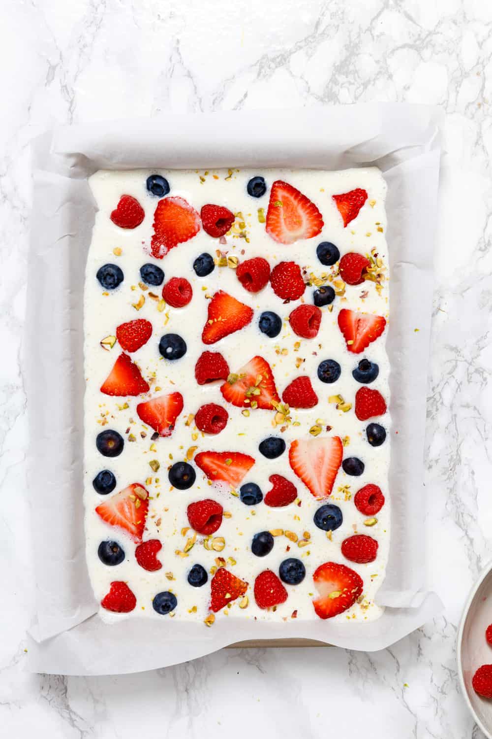 an overhead shot of yogurt, berries and nuts layered in a sheet pan lined with parchment paper before freezing