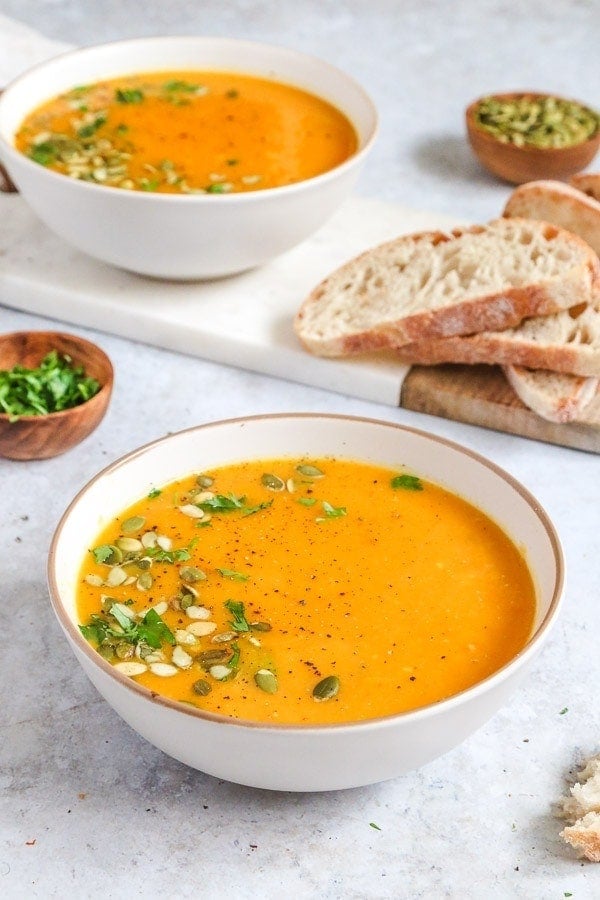 A bowl of butternut squash soup topped with sunflower seeds and chopped parsley with crusty bread on a board