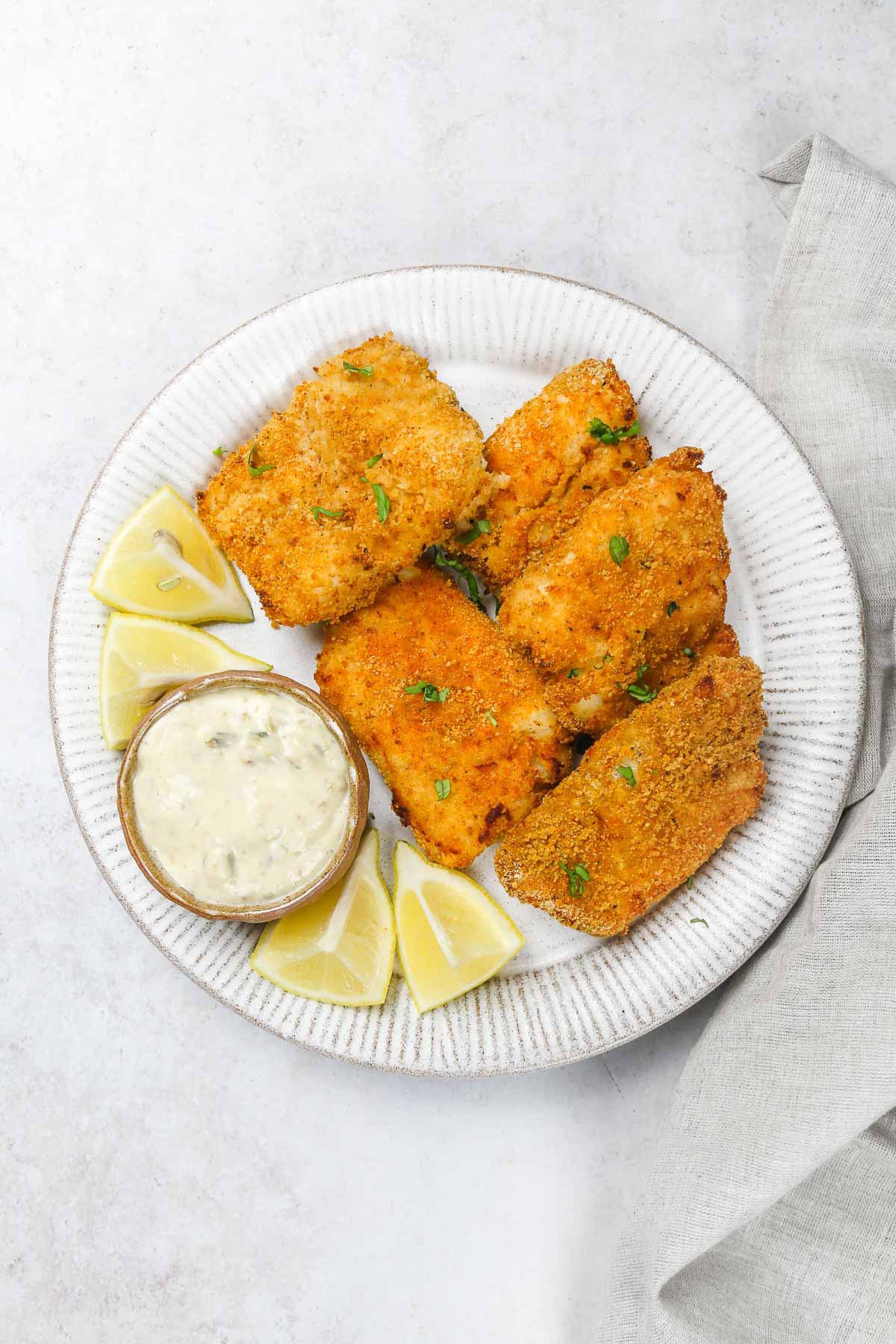 A white plate with 6 pieces of crispy air fried fish, lemon wedges, and tartar sauce