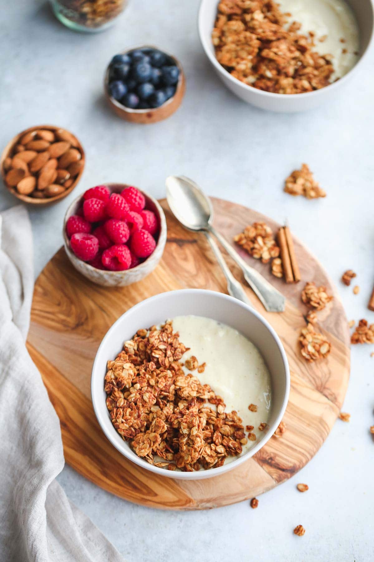 Granola and yogurt in a bowl on a wooden board, with small bowls around it filled with fruit and nuts