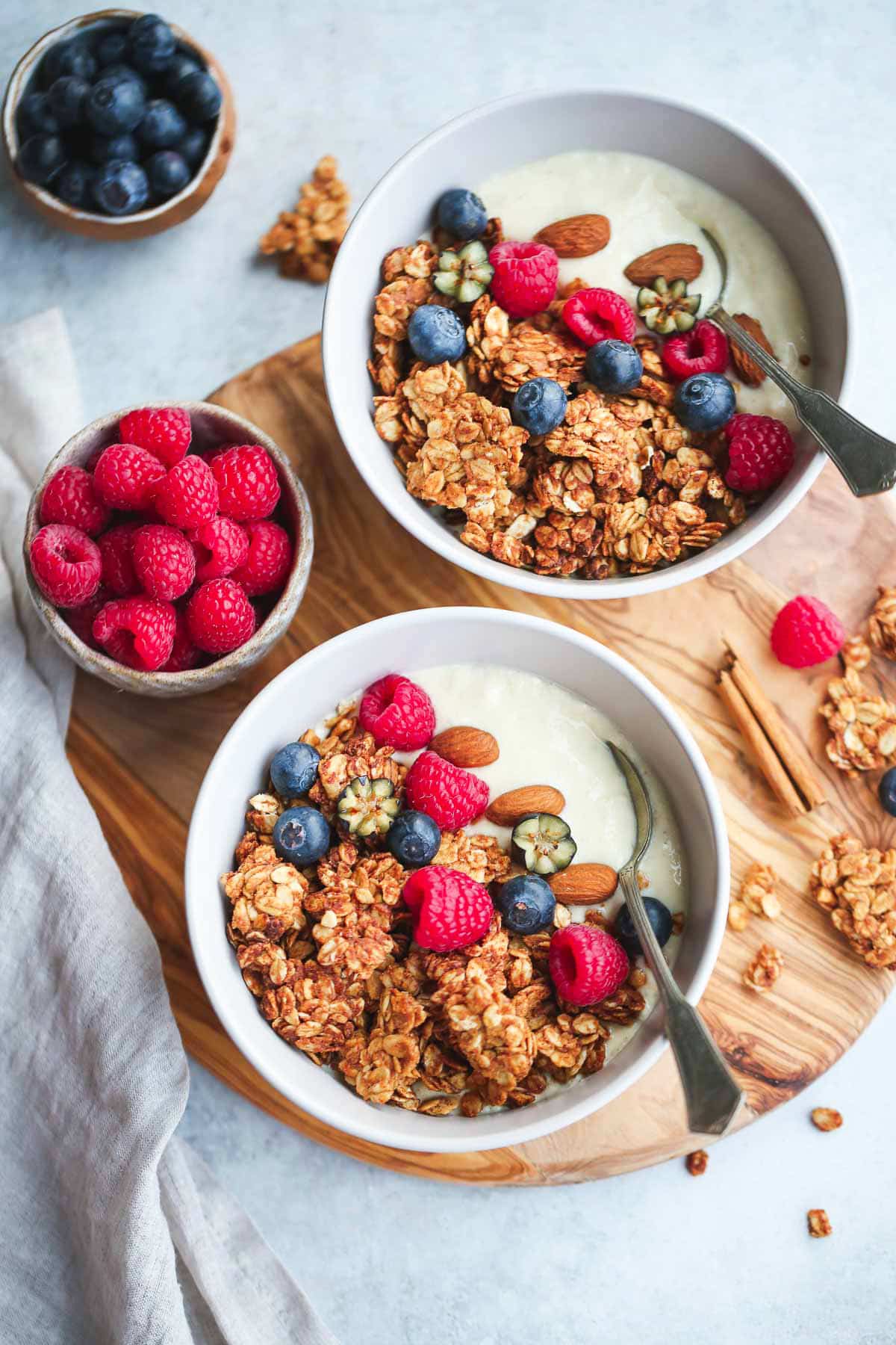 2 bowls of granola with yogurt and fresh berries on a wooden board, and a tea towel on the side
