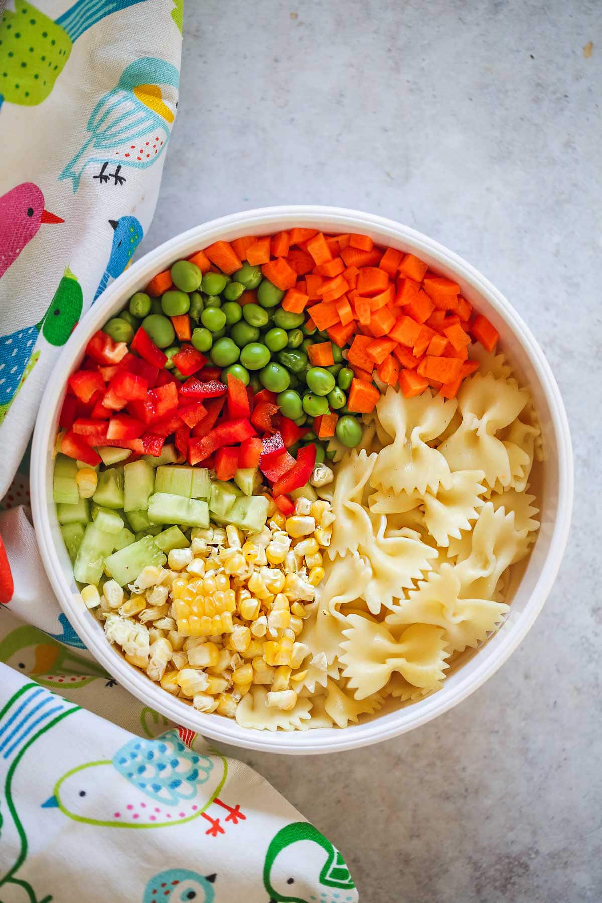 The ingredients of the pasta salad layered in a white bowl, and a bird pattern kitchen towel around it