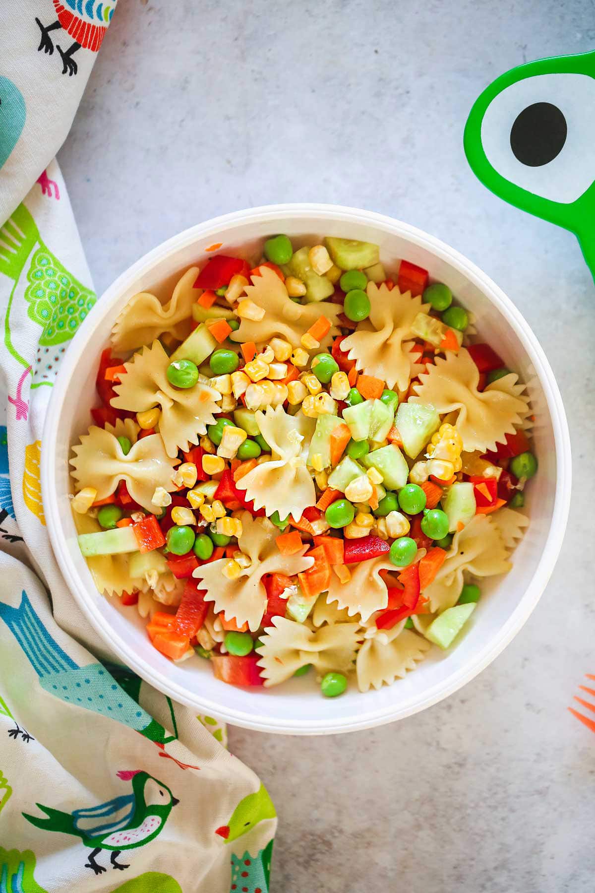 Pasta salad with sweet corn, peas, carrots, and sweet bell peppers in a white bowl and a cute bird pattern kitchen towel