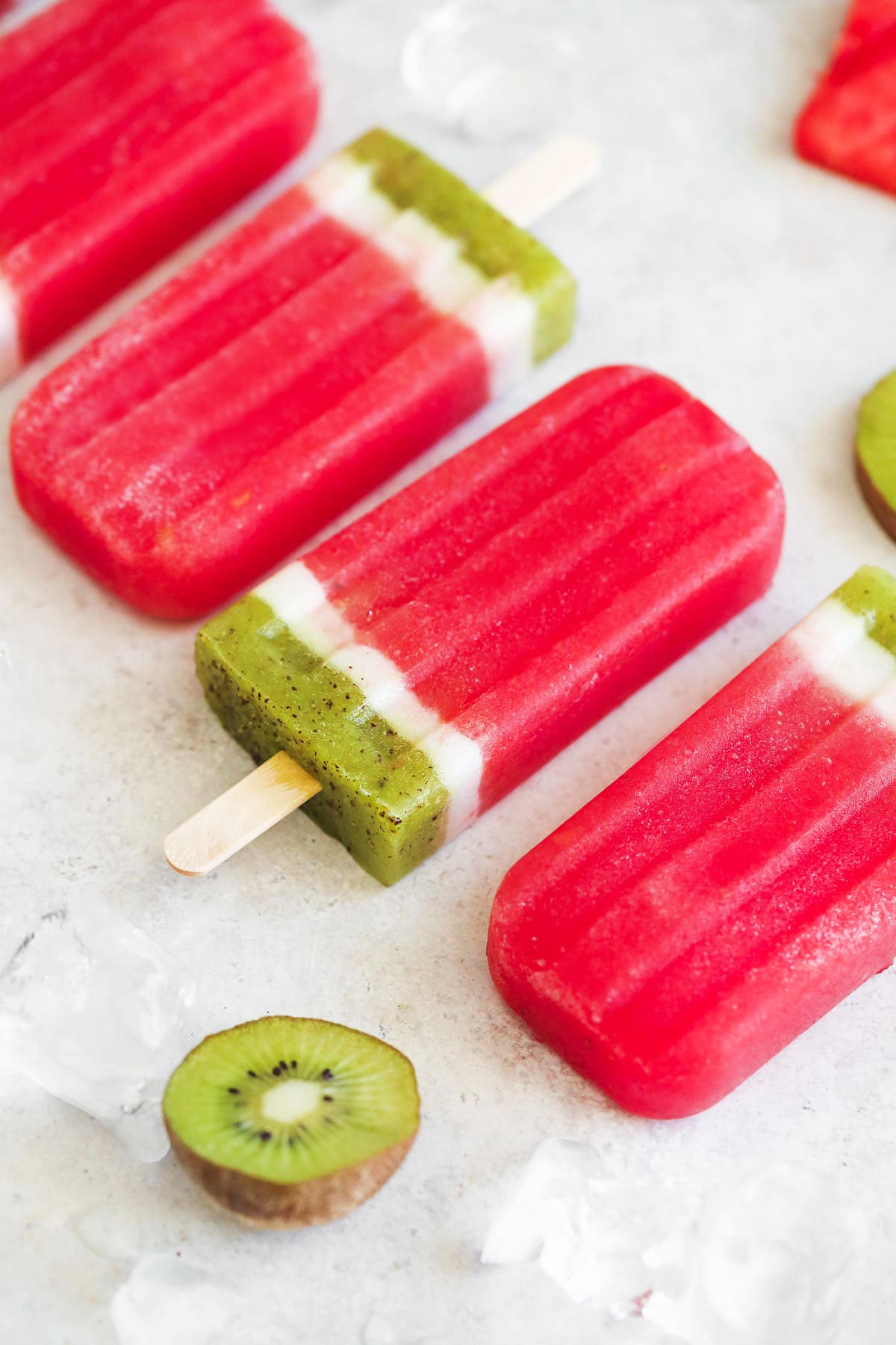 Watermelon popsicles placed on a counter with ice cubes, and a fresh halved kiwi