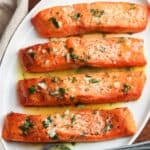 Four cooked salmon fillets on a white plate, topped with garlic butter and fresh herbs, served with lemon on the side.