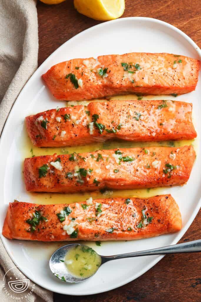 Four cooked salmon fillets on a white plate, topped with garlic butter and fresh herbs, served with lemon on the side.