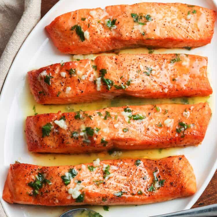 Four cooked salmon fillets on a white plate, topped with garlic butter and fresh herbs, served with lemon on the side.