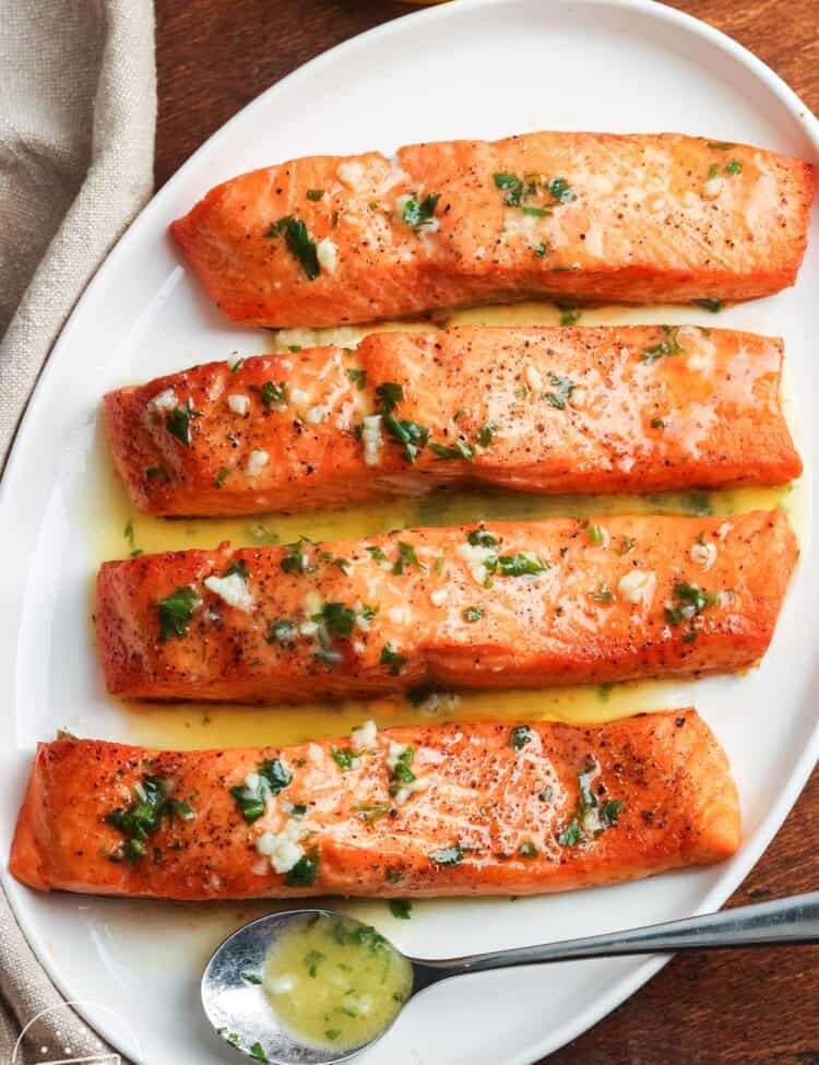 Four cooked salmon fillets on a white plate, topped with garlic butter and fresh herbs, served with lemon on the side.