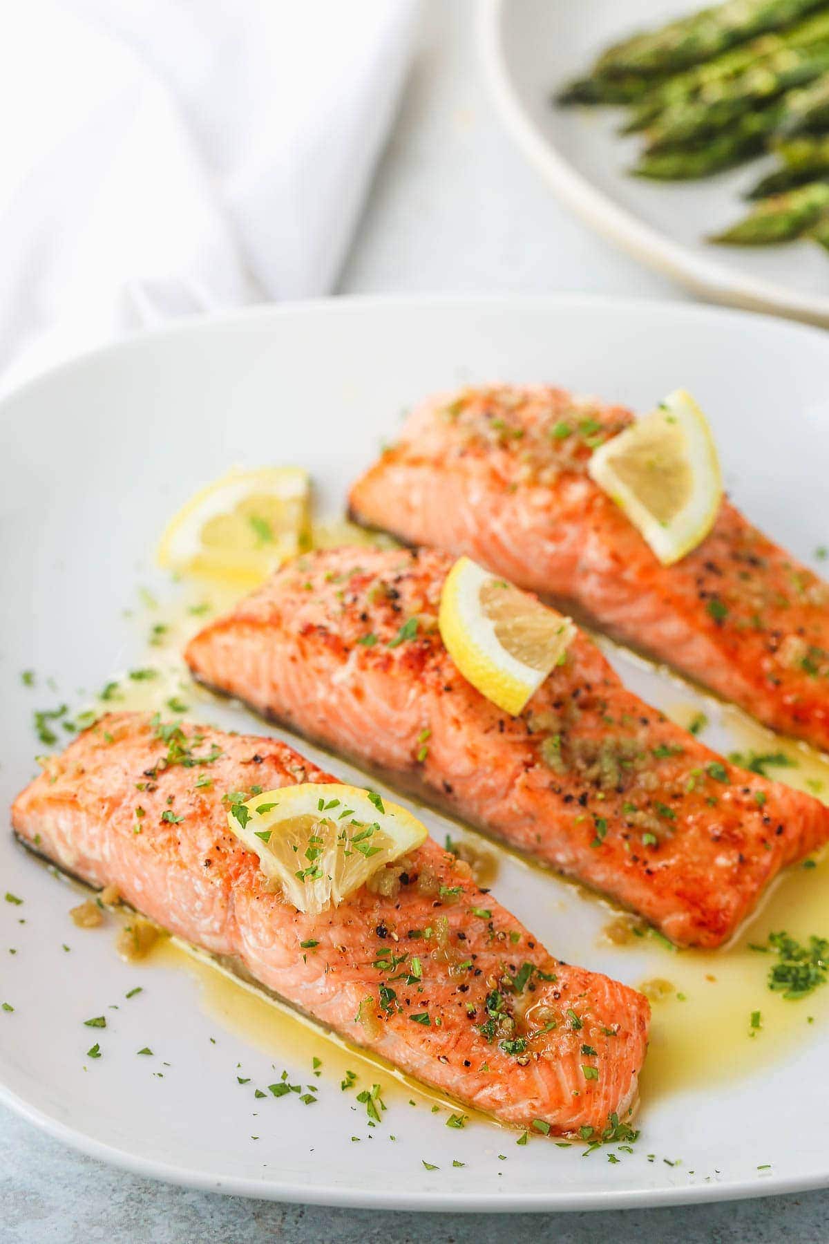 Three air fryer salmon fillets topped with a slice of lemon and chopped parsley on a white plate.