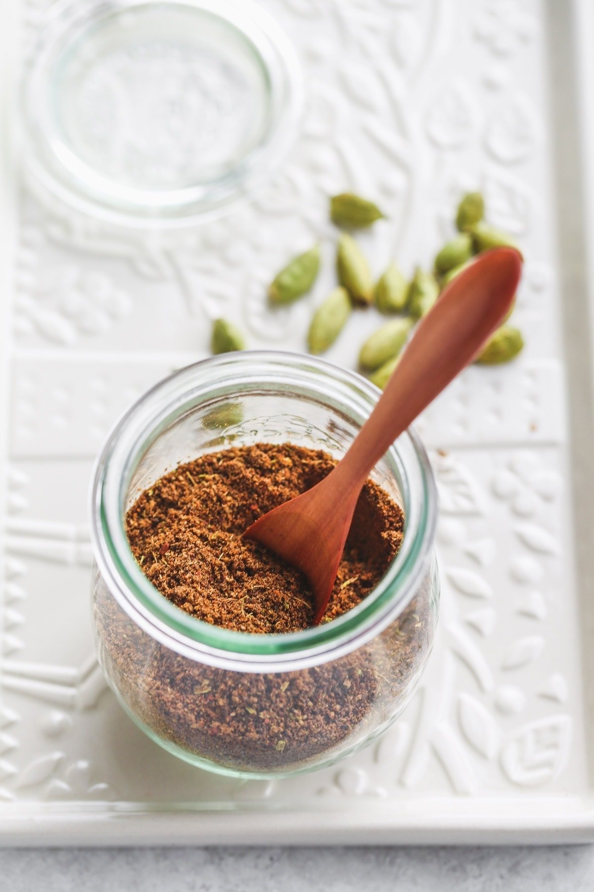 A Weck jar filled with baharat spice blend and a wooden teaspoon, cardamom pods on a white tray.