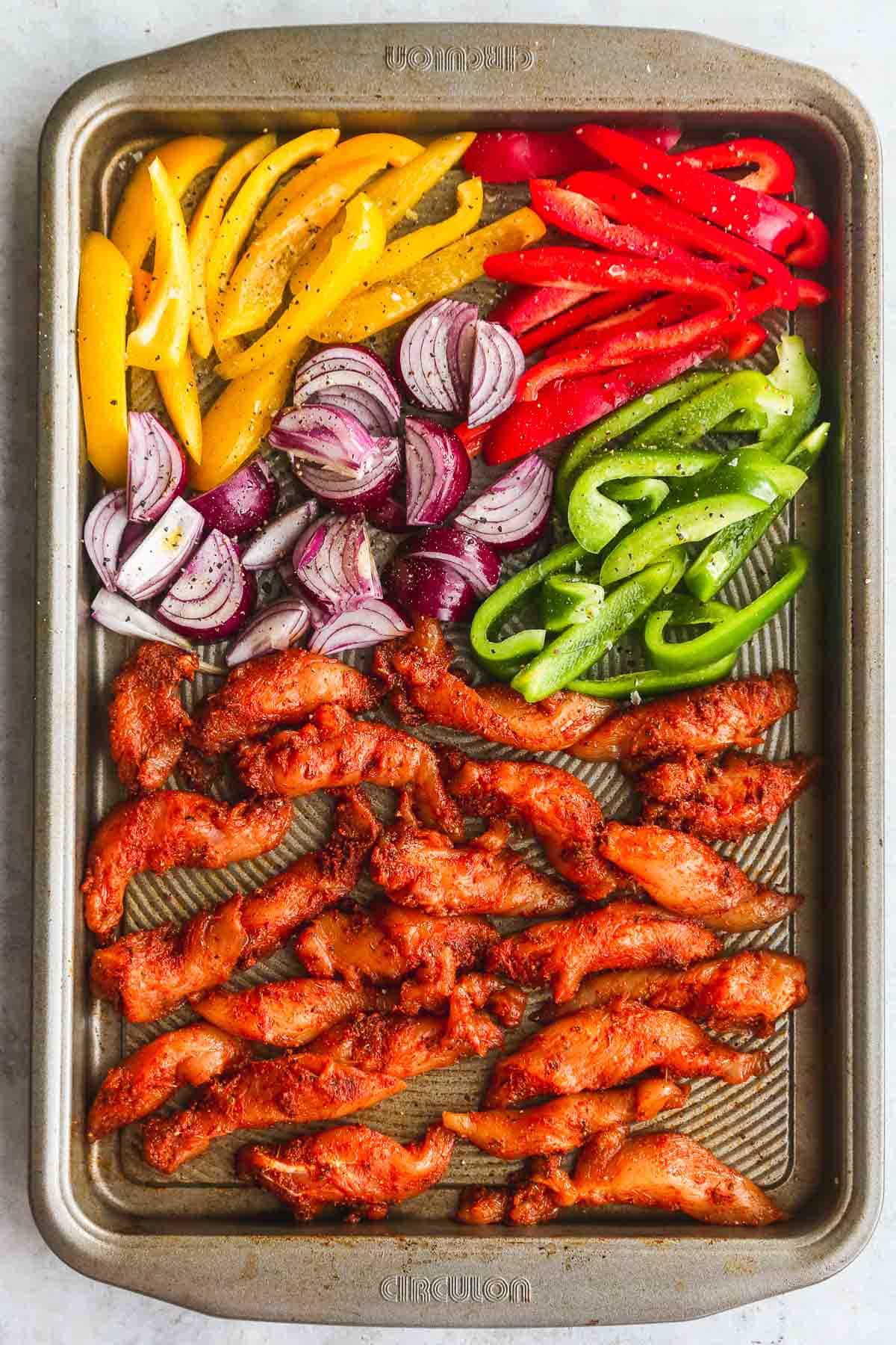 Raw seasoned chicken fajitas and vegetables on a sheet pan.
