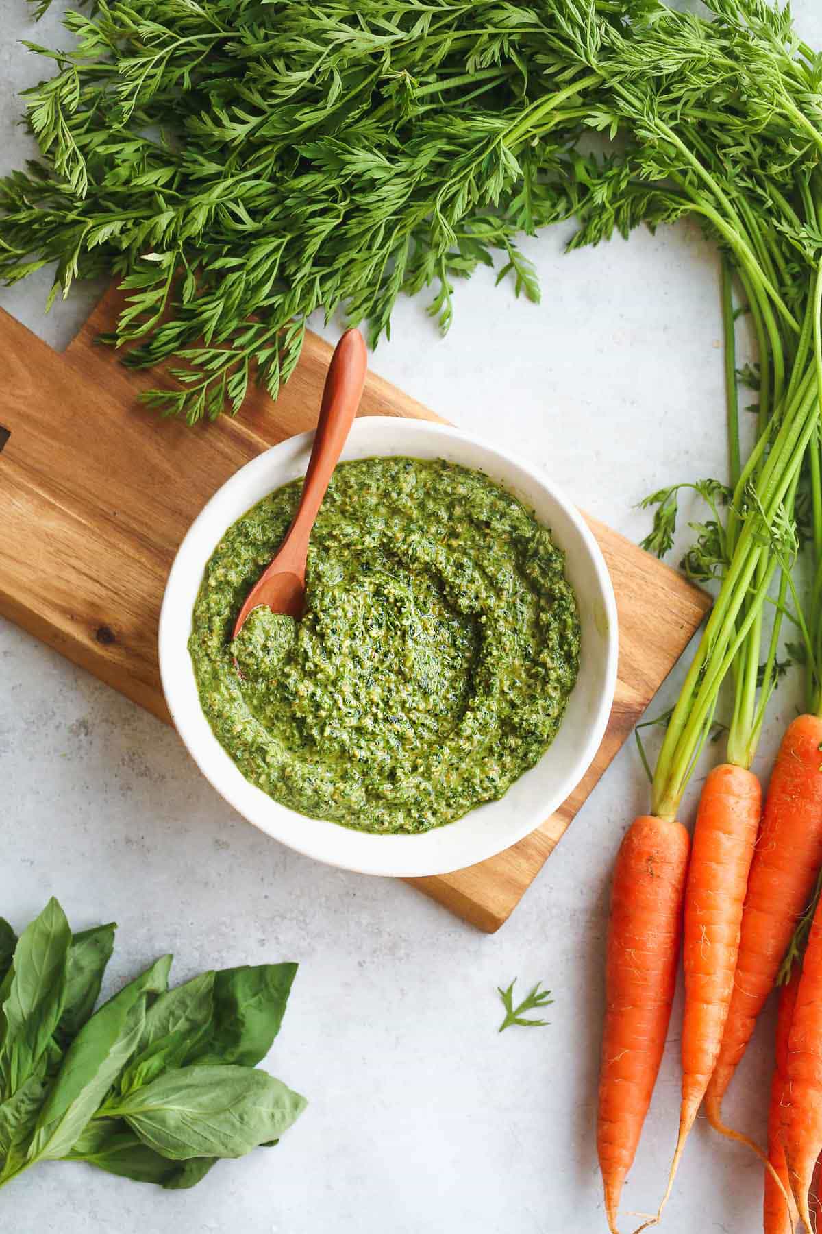 Carrot Top Pesto in a small white bowl on a wooden board. Carrots, basil leaves are laying on the side of the bowl.