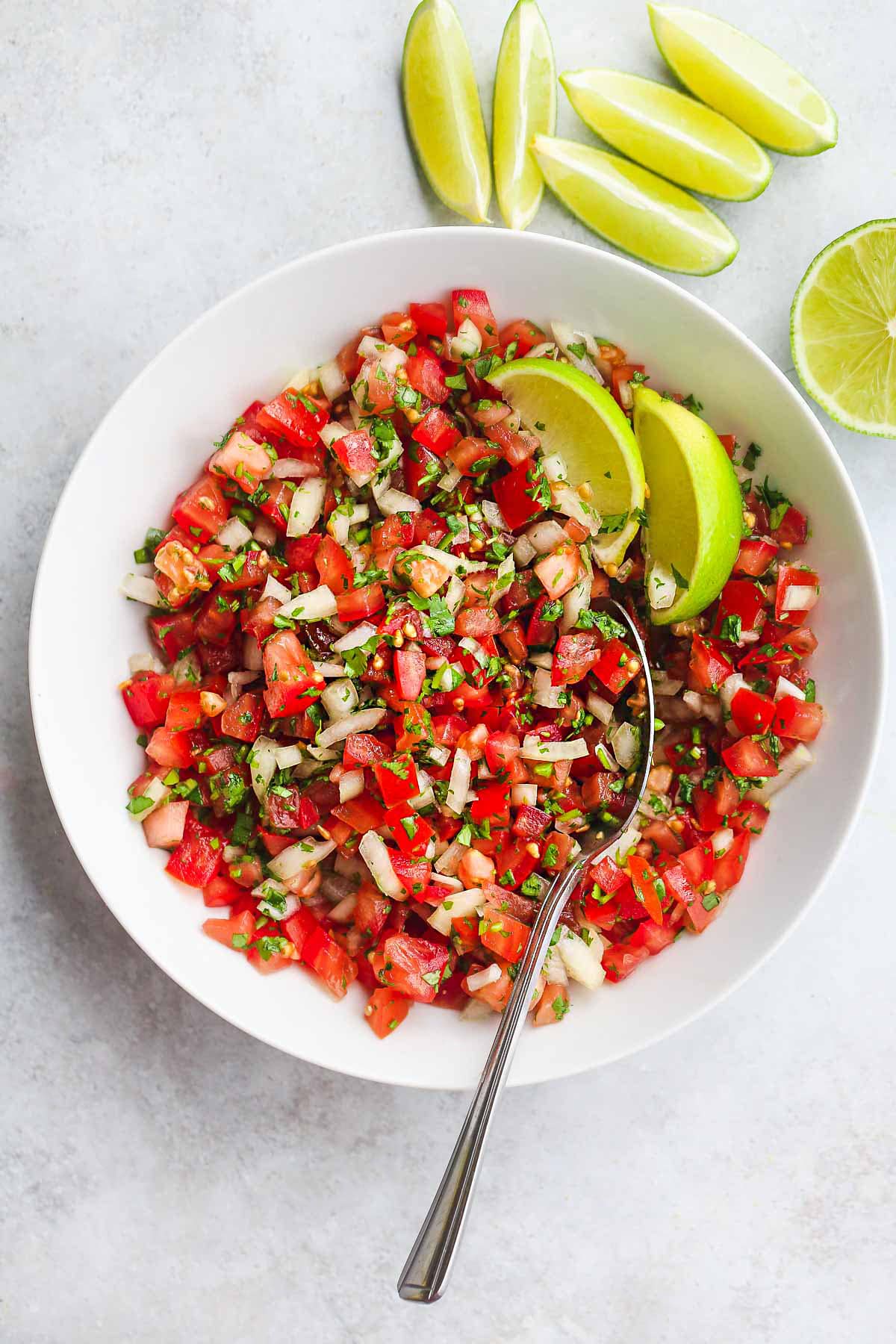 A white bowl with Pico De Gallo Salsa, a serving spoon and lime wedges
