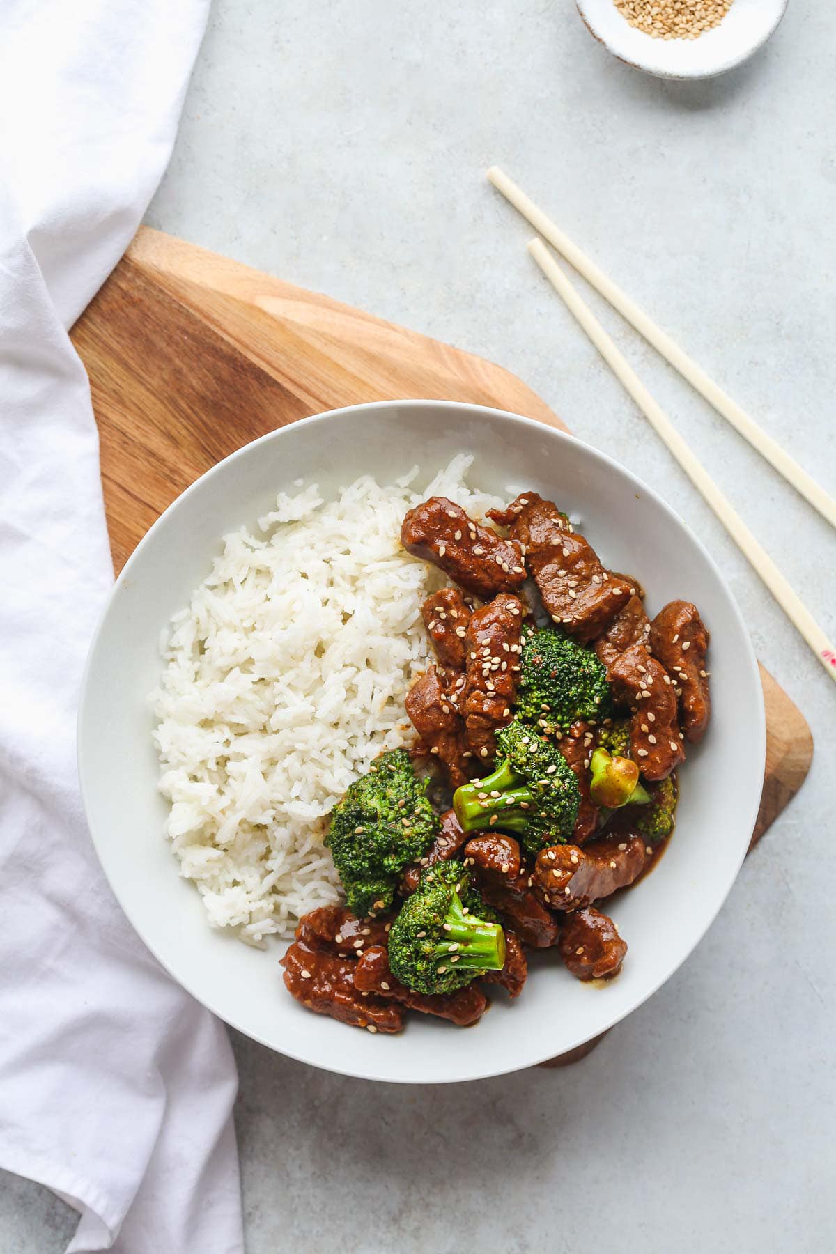 A white bowl with white rice, and beef and broccoli garnished with toasted sesame seeds. With chop sticks on the side