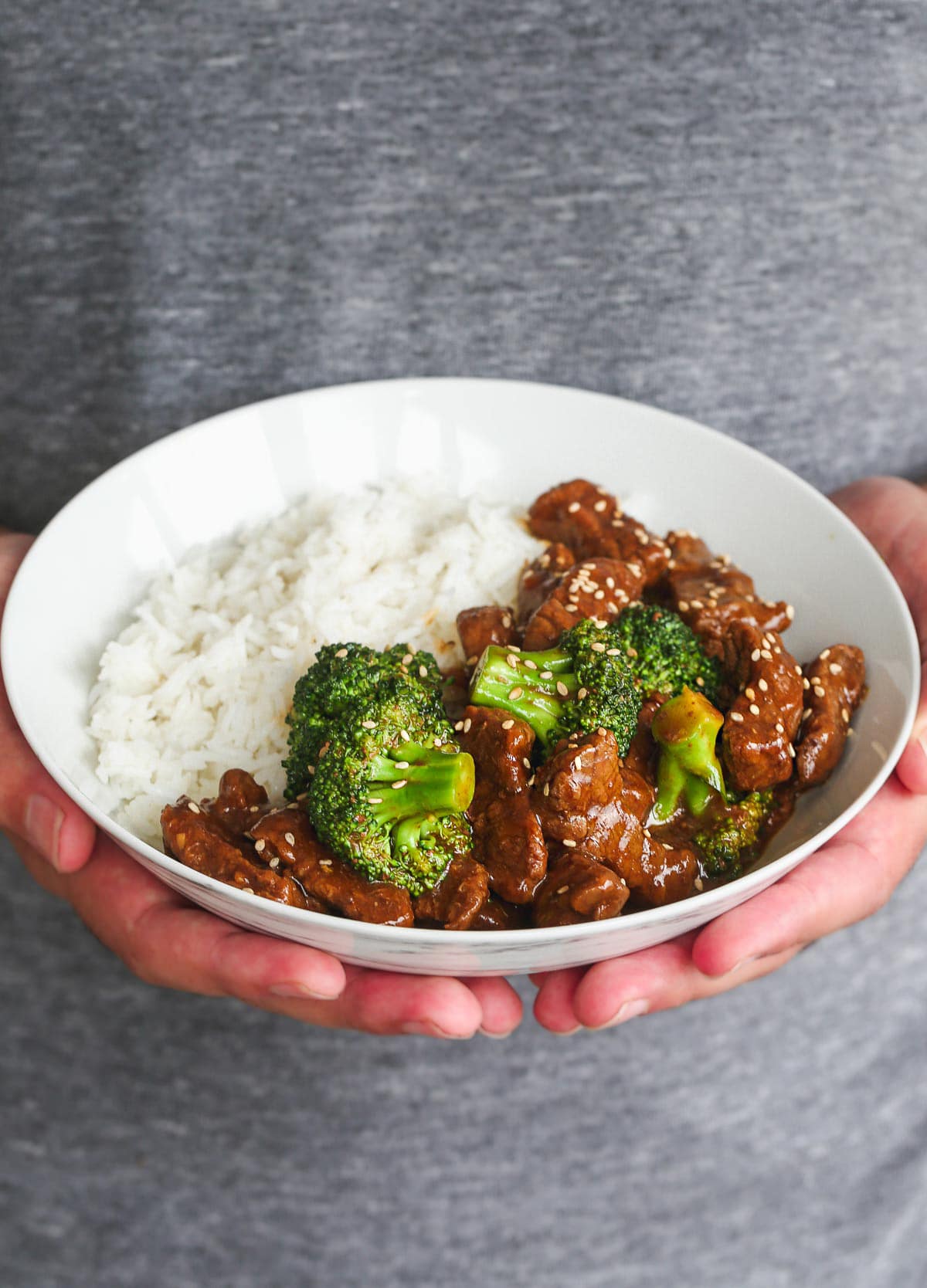 Someone holding a white bowl of beef and broccoli with rice in their hands