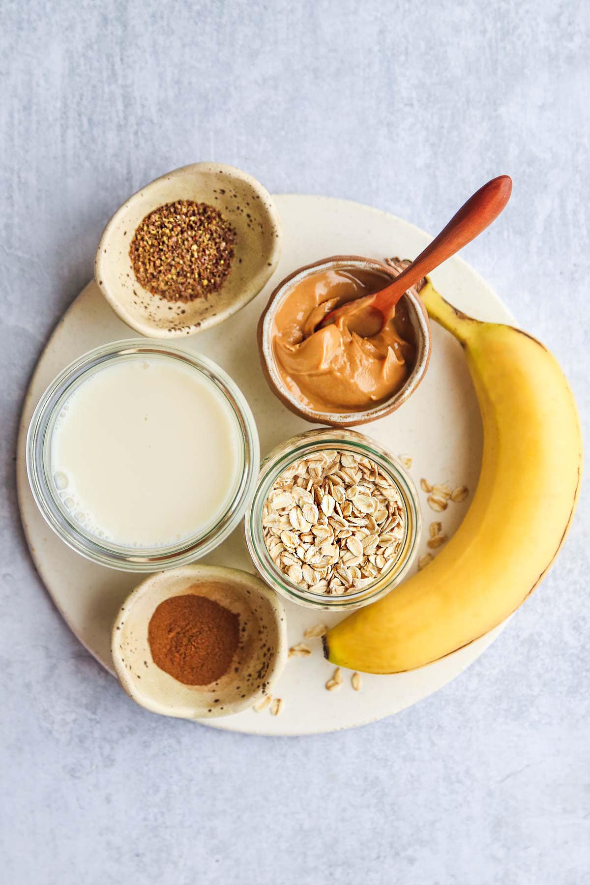 Banana oatmeal smoothie ingredients laid on a tray including a banana, peanut butter, cinnamon, almond milk, ground flaxseed, and oats
