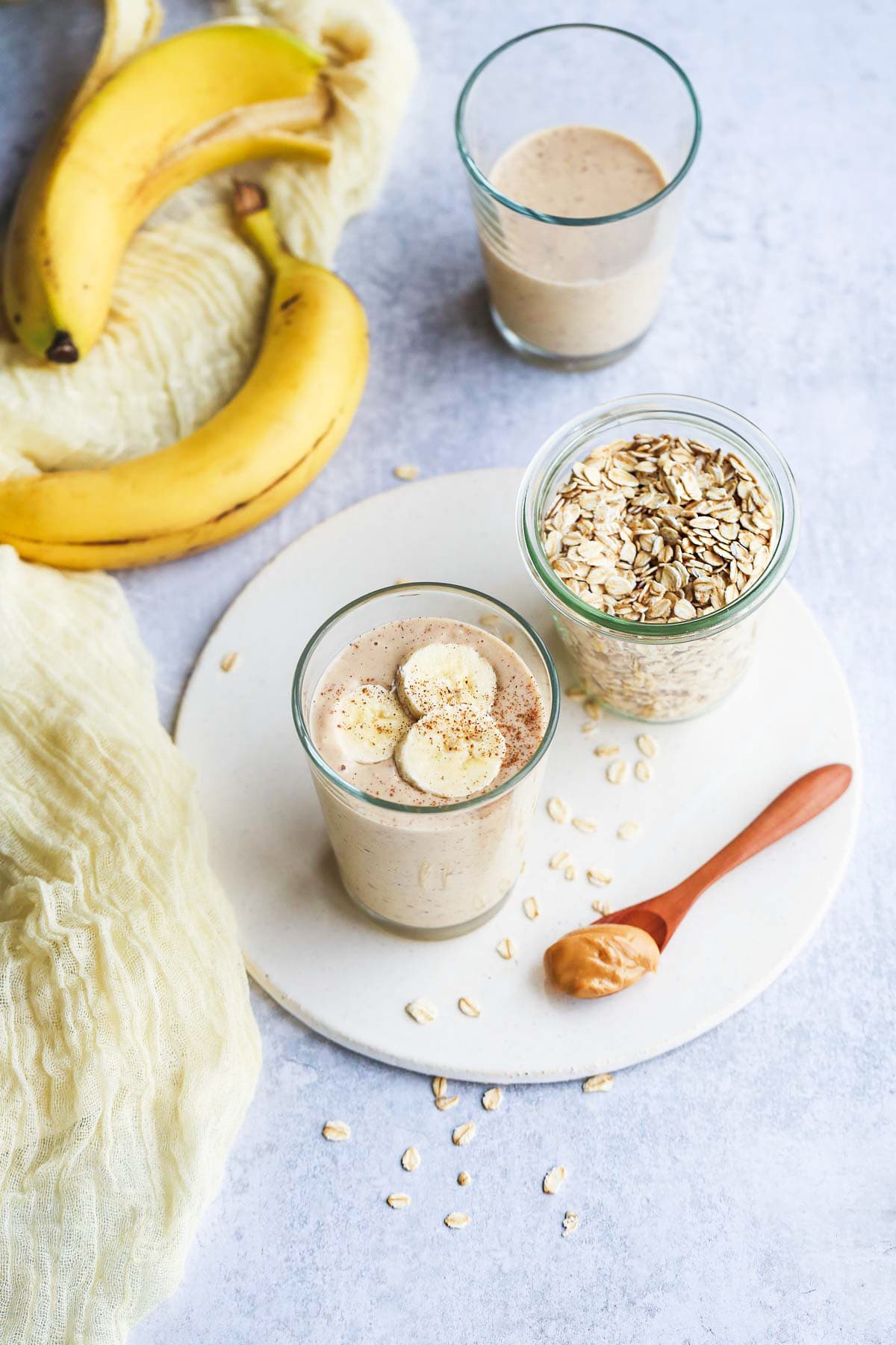 A glass of banana oatmeal smoothie along with a jar of rolled oats on a white tray