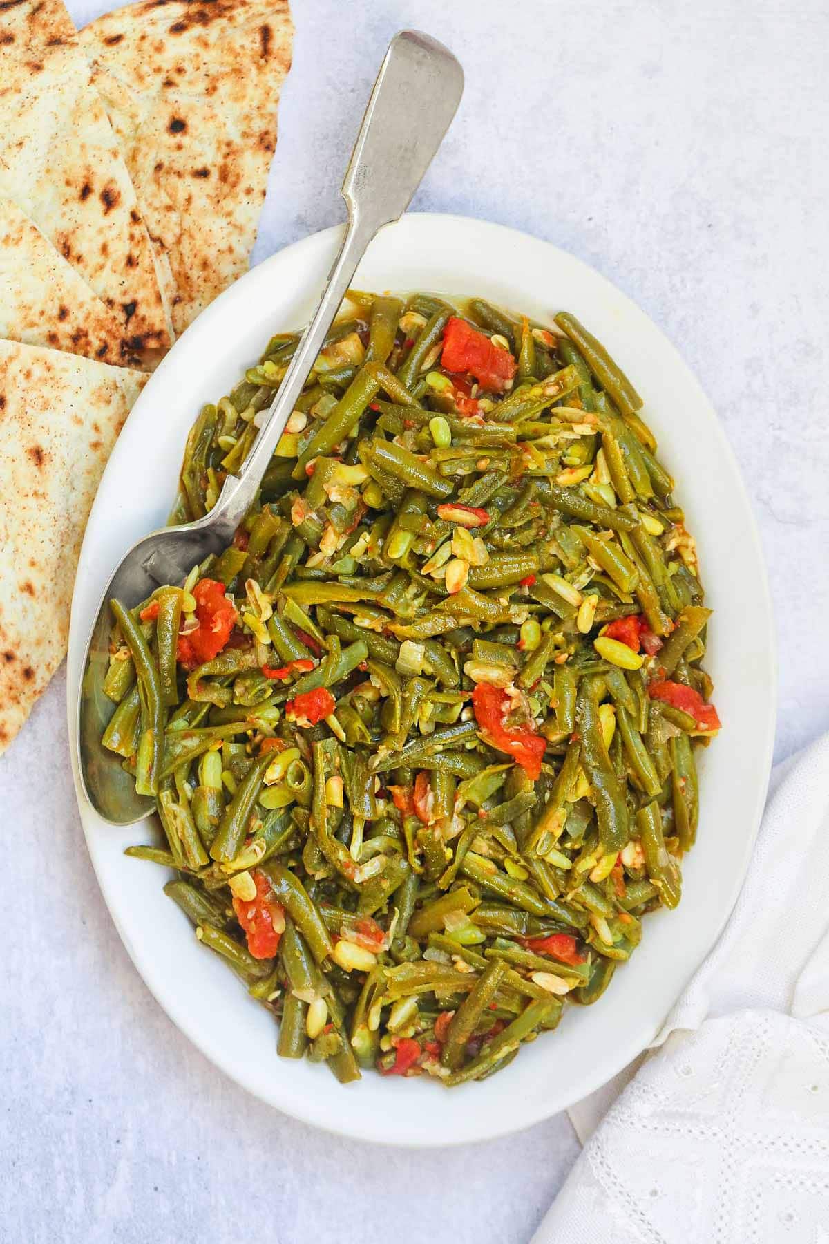 Stewed green beans on a large serving plate and a serving spoon, and pita bread on the side.