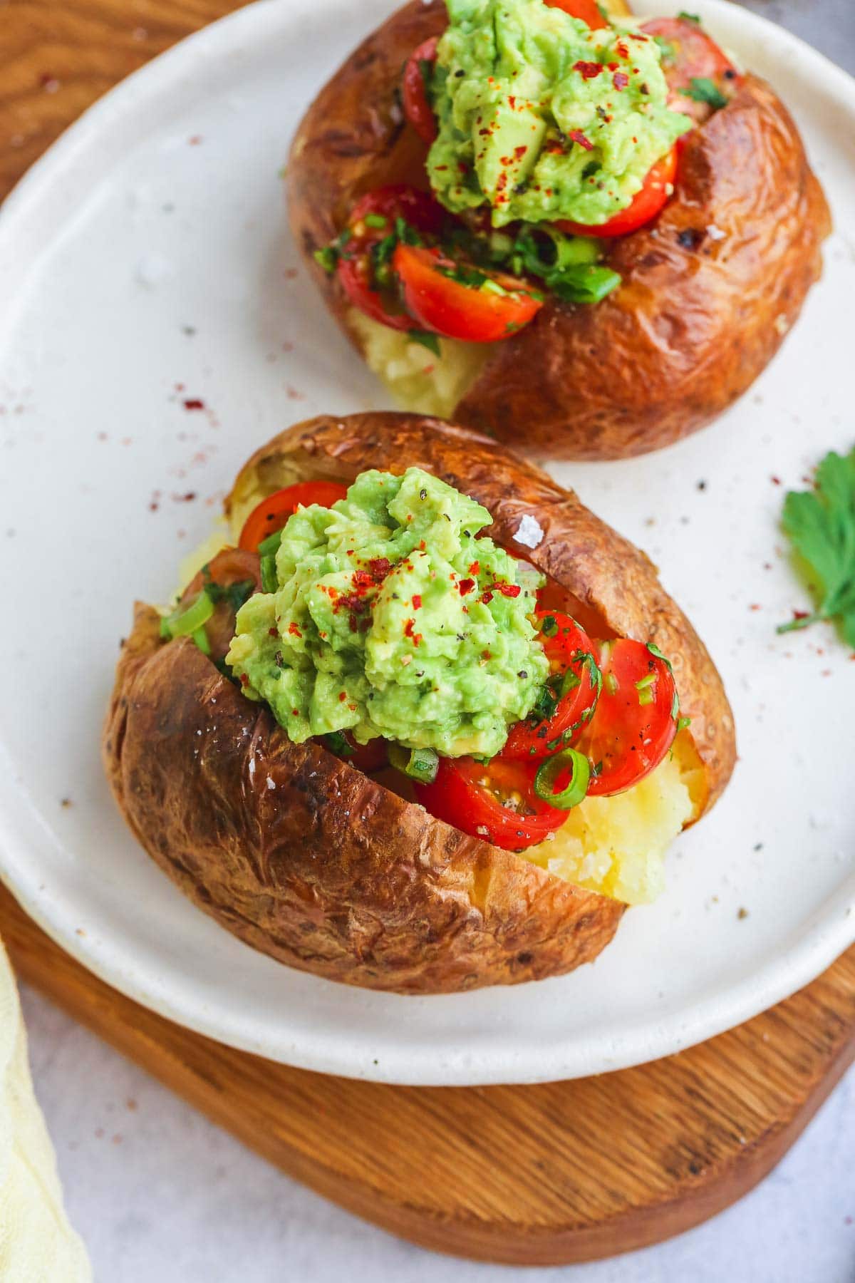 Air Fryer baked potato topped with tomato salad and smashed avocado on a white plate and a wooden cutting board.