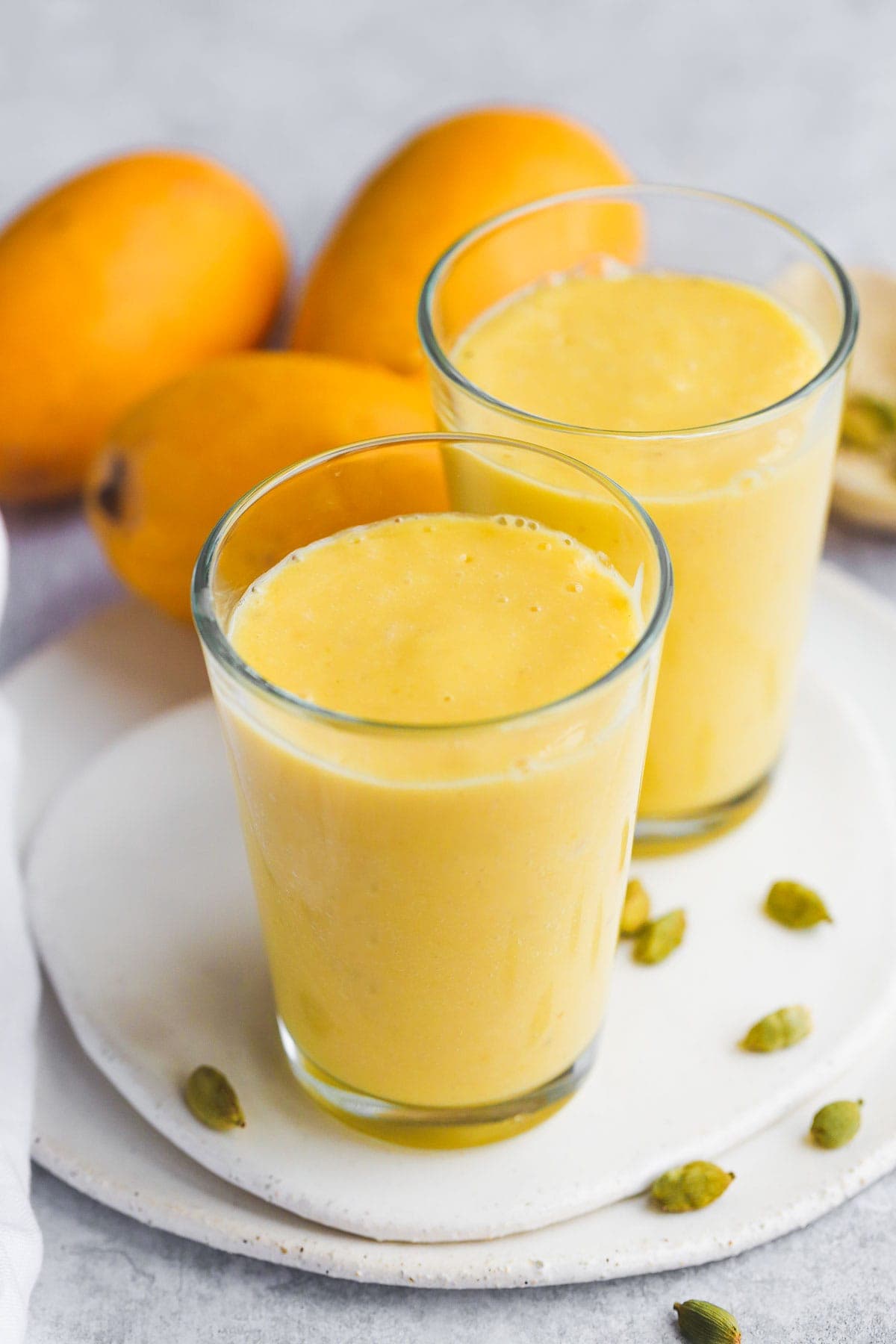 2 glasses with mango lassi on ceramic white tray, 3 mangoes in the background, and some green cardamom pods