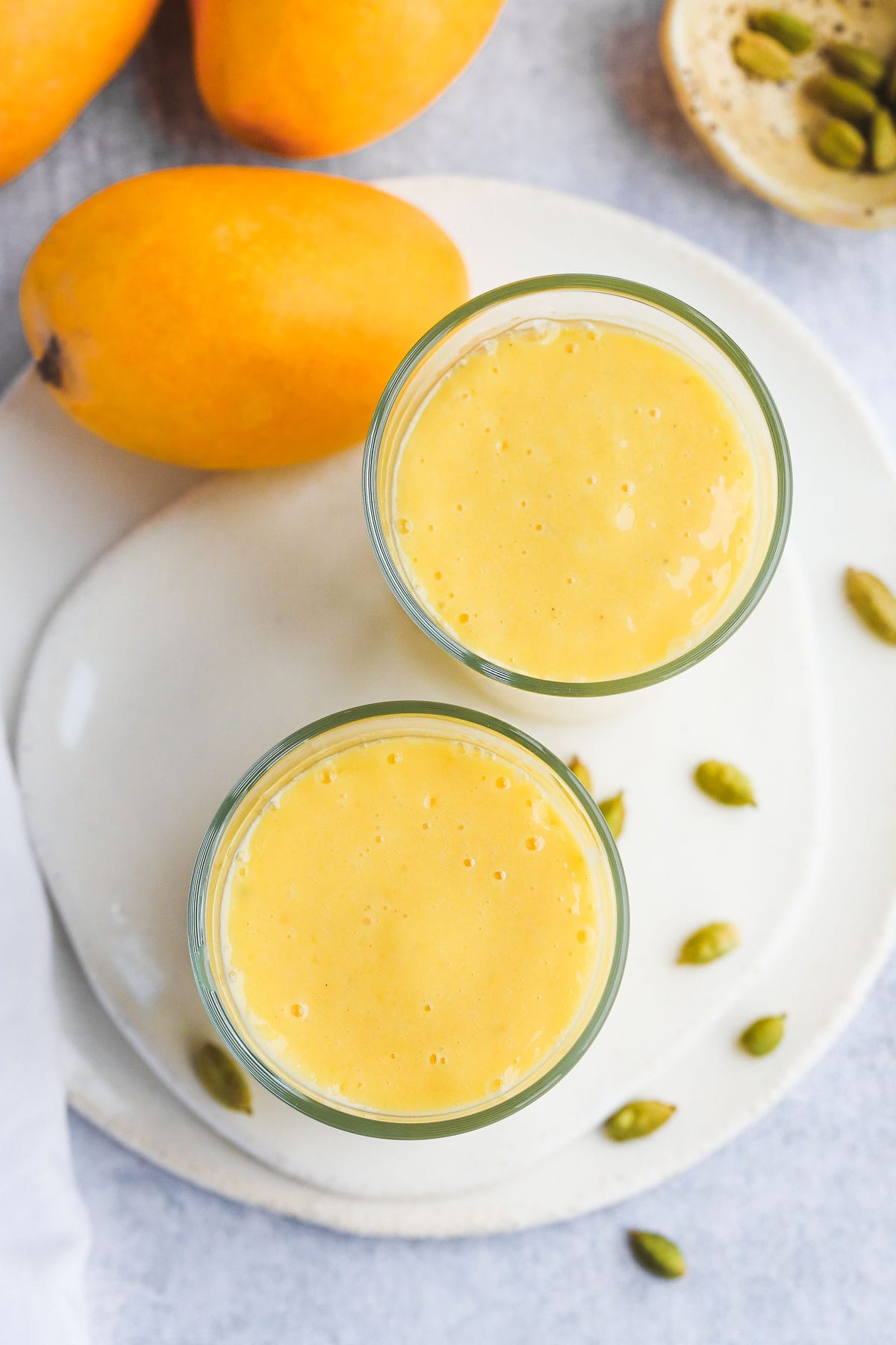 Overhead shot of 2 glasses with mango lassi on ceramic white tray, 3 mangoes in the background, and some green cardamom pods