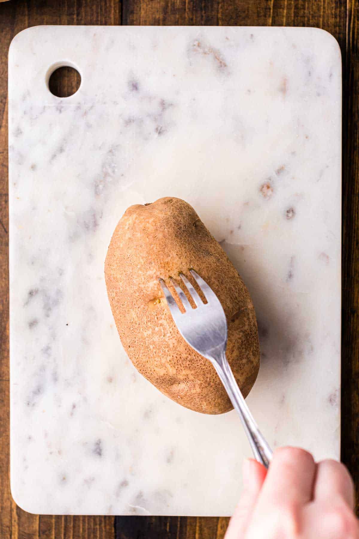 Russet potato being pierced with a fork on a cutting board before air frying