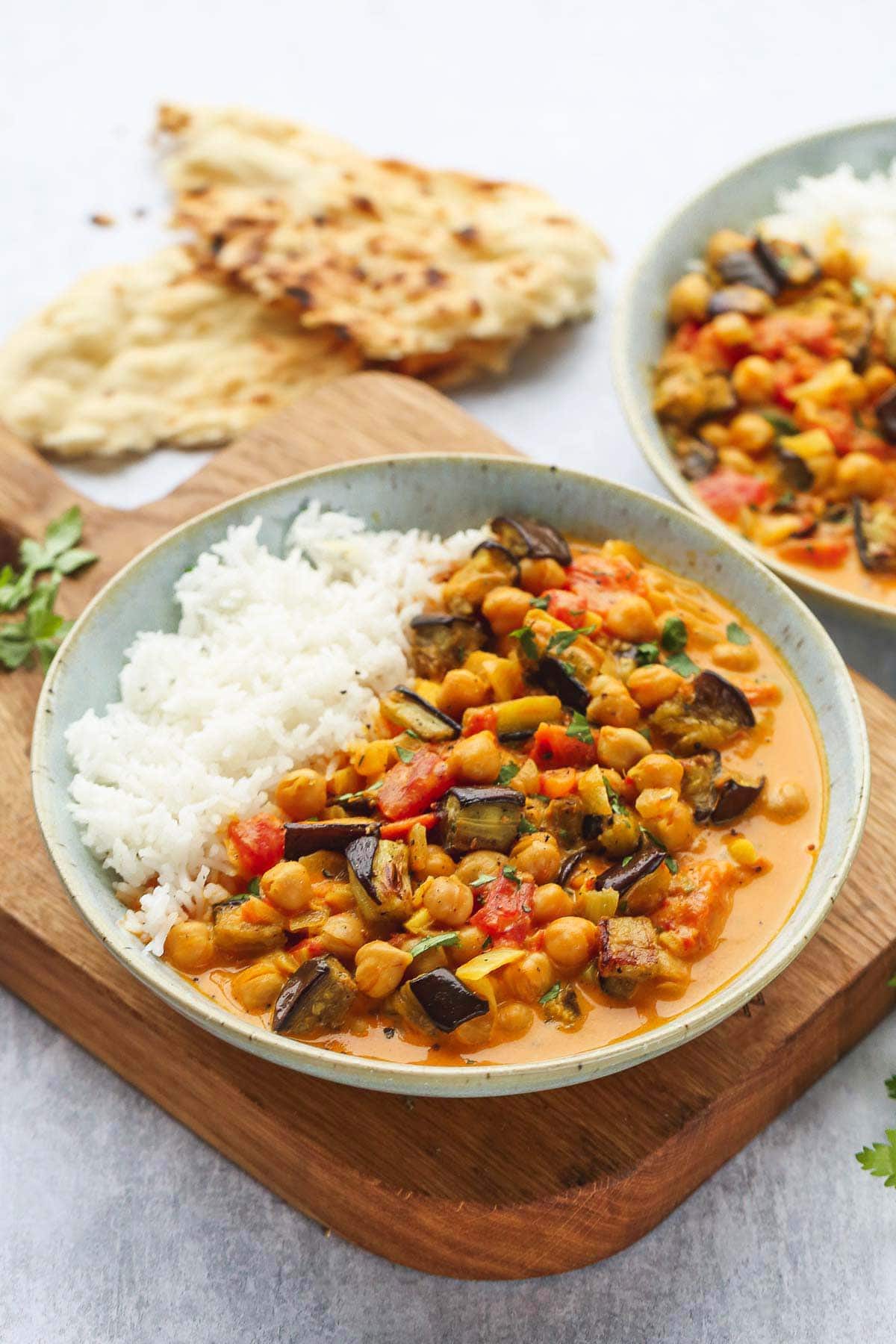 A bowl of Aubergine and Chickpea Curry served with white basmati rice over a wooden board.