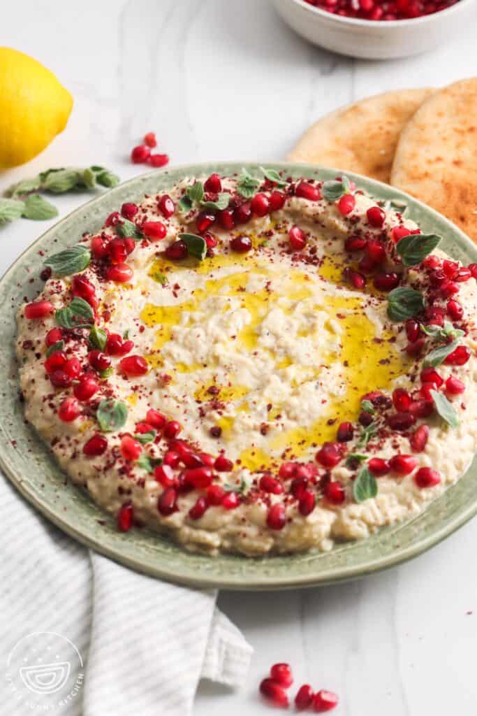 A green ceramic plate filled with creamy mutabal, drizzled with golden olive oil, and garnished with bright red pomegranate seeds, sumac, and fresh oregano leaves. Soft pita bread is placed on the side.