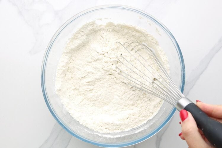 A hand holding a whisk in a bowl of flour and other dry ingredients