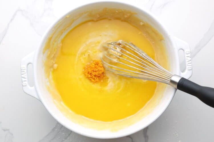 Orange juice and zest added to cake batter before mixing in a white bowl, and a whisk.