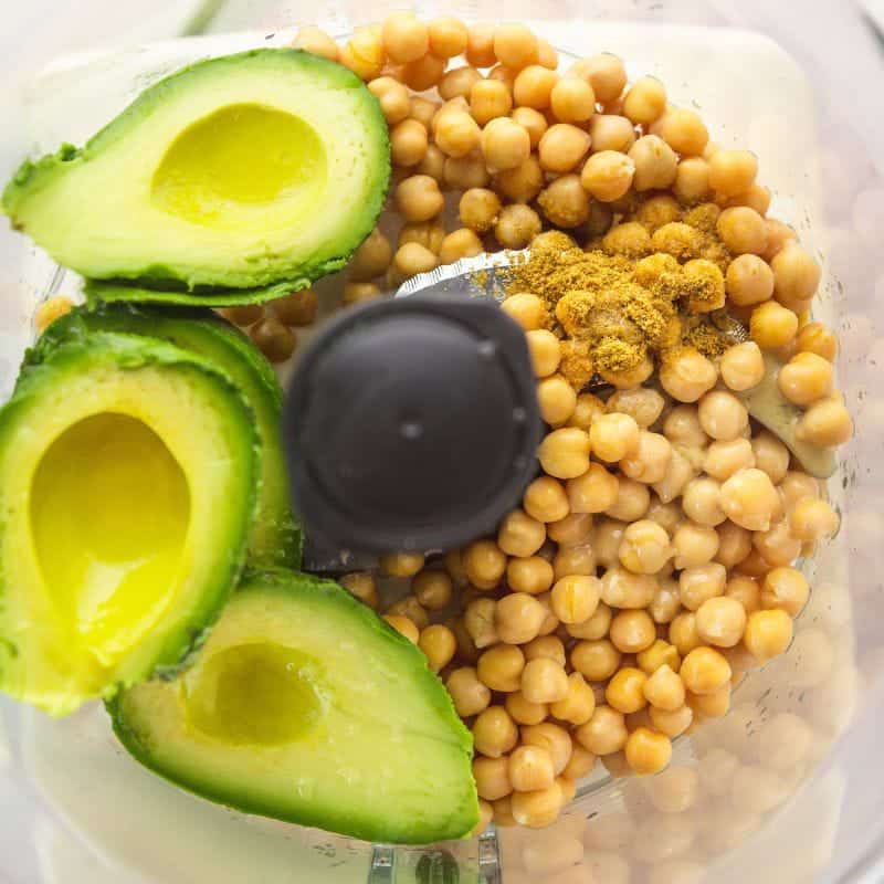 Chickpeas, ripe avocado, and spices in a food processor ready to be blended into hummus